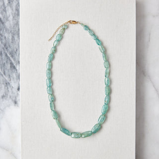 A beaded necklace made of light aqua-blue oval beads with a small gold clasp, displayed on a white textured card over a gray and white marble surface.