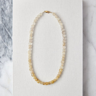 A beaded necklace made of small translucent pale yellow and cream faceted beads with a gold clasp, displayed on a white textured card over a gray-and-white marble surface.