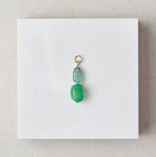 Jewelry pendant with green gemstone on a white card, placed on a beige square with marble background
