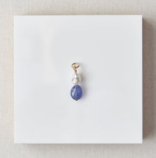 Jewelry pendant with blue stone on a white card, placed on a beige square with marble background