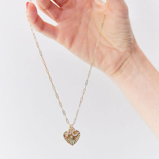 Hand holding a gold necklace with a heart pendant and two dazzle letters against a light background