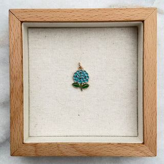 A small enamel flower-shaped pin with blue and green detailing on a gold loop, displayed inside a square wooden shadow box with a beige fabric lining, set on a white and gray marble surface.