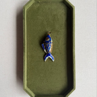 A small fish-shaped enamel pendant with blue, white, and gold detailing, displayed inside an olive green velvet jewelry box on a light beige surface.