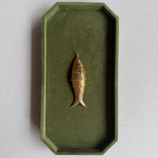A small gold fish-shaped brooch or pin displayed in the center of an olive green octagonal velvet jewelry box, set against a light beige linen background.