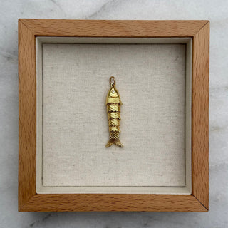 A small gold fish-shaped pendant or charm displayed inside a square wooden shadow box with a beige fabric lining, placed on a white marble surface.
