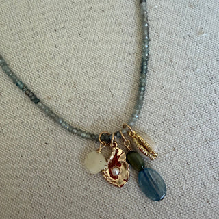 A close-up of a beaded necklace with a gold-toned charm setting featuring a white stone, a textured gold shell-shaped charm with a red center, a small green bead, and a blue faceted bead, displayed on a beige fabric background.