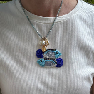 A close-up of a person wearing a white ribbed t-shirt and a detailed beaded necklace featuring two fish and small shell-like beads.