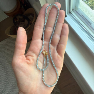 A delicate beaded necklace with light blue-gray beads and a small gold clasp, held in the palm of a hand near a window.