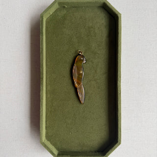 A small polished brown and amber bird-shaped pendant or charm displayed inside an olive green octagonal jewelry box on a light neutral surface.