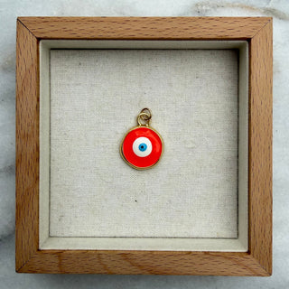 A small red evil eye pendant with a white center and gold bail, displayed inside a square wooden shadow box on a light marble surface.