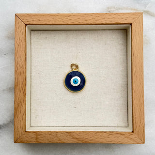 A small evil eye pendant with a blue and white circular design and a gold bail, displayed inside a square light wood shadow box on a white and gray marble surface.
