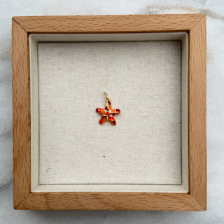 A small orange-red starfish-shaped pendant displayed inside a square light wood shadow box with a cream fabric lining, set on a white marble surface.