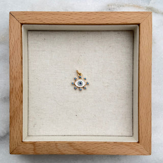 A small gold charm with a blue and white eye design sits in the center of an open square wooden jewelry box on a white and gray marble surface.