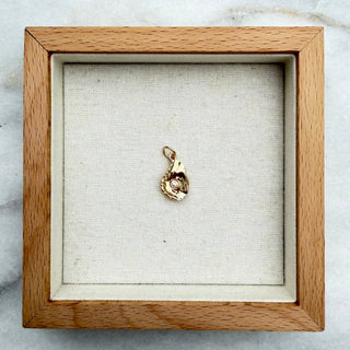 A small gold pendant or charm displayed inside a square wooden shadow box with a cream fabric lining, placed on a white and gray marble surface.