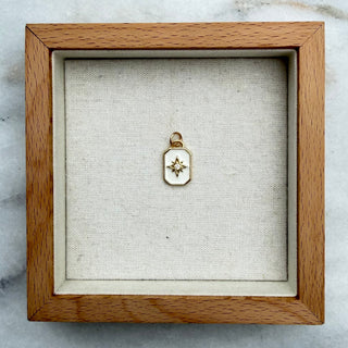 A small gold pendant with a star-like design inside a square wooden shadow box display, placed on a white and gray marble surface.