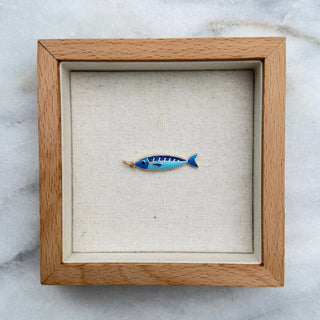 A small blue and white fish-shaped pendant or charm displayed inside a square light wood shadow box with a cream fabric lining, placed on a white marble surface.