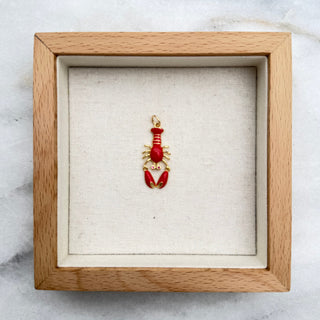 A small lobster-shaped pendant in red and gold, displayed inside a light wood shadow box with a cream fabric lining, set on a white marble surface.