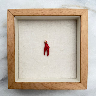 A miniature red lobster claw pendant displayed inside a square light wood shadow box with a cream fabric lining, set against a white marble background.