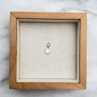 A small mother of pearl fish pendant, displayed inside a square light wood shadow box on a white and gray marble surface.