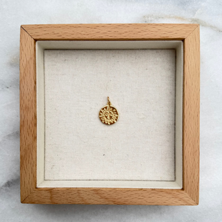 A small gold pendant with a sunburst or starburst design, displayed inside a square light wood shadow box with a cream fabric lining, set on a white marble surface.