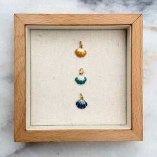 Three small shell-shaped pendants displayed in a light wood shadow box on a white and gray marble surface.