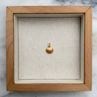 A small gold pendant displayed inside a square wooden shadow box with a cream fabric lining, set on a light gray marble surface.