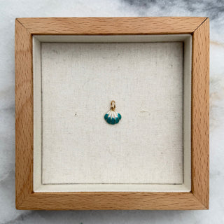 A small gold pendant with a green and white marbled bead charm, displayed inside a square light wood shadow box on a white and gray marble surface.