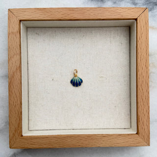 A small shell-shaped pendant with blue and green striped enamel and a gold-tone top loop, displayed inside a square light wood shadow box on a white marble surface.