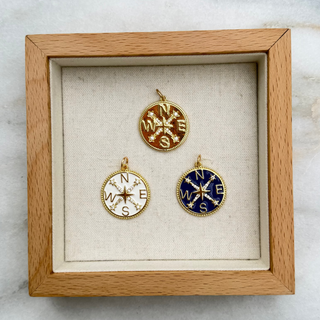 Three small compass pendant charms in gold, white, and blue enamel are displayed inside a square light wood shadow box on a white marble surface.