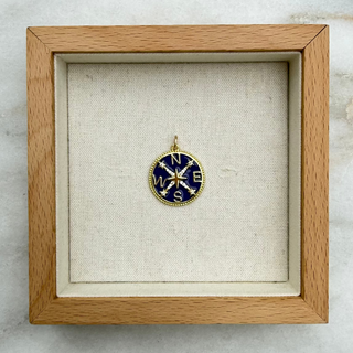 A small round compass pendant with a gold-toned frame and dark blue enamel face, displayed inside a square light wood shadow box on a marble surface.
