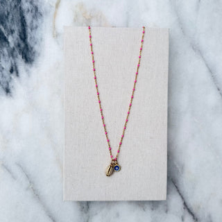 A delicate necklace with a small gold pendant featuring a blue eye motif, displayed on a textured beige card over a white and gray marble surface.