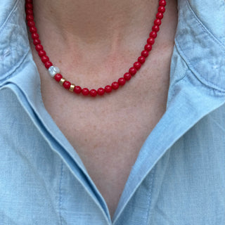 A close-up of a person wearing a red beaded necklace with a small white and gold accent bead, paired with a light blue button-up shirt.