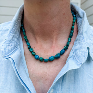 A close-up of a person wearing a chunky turquoise and teal beaded necklace with a light blue linen shirt.
