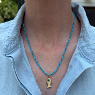 A close-up of a person wearing a turquoise beaded necklace with a gold fish-shaped pendant featuring a small blue stone accent.