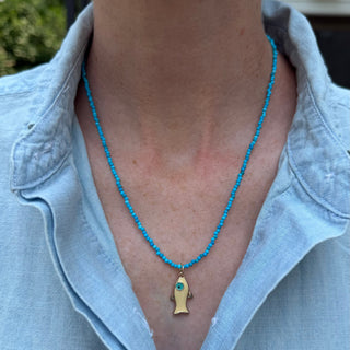 A close-up of a person wearing a turquoise beaded necklace with a gold fish-shaped pendant featuring a small blue stone accent.