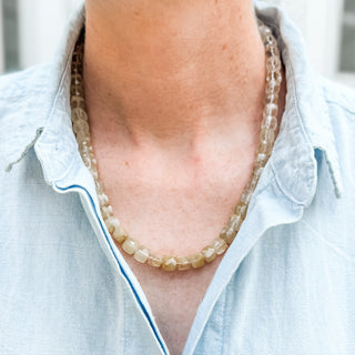A close-up of a person wearing a double-strand beaded necklace made of translucent light brown and beige stone beads, paired with a light blue linen button-up shirt.