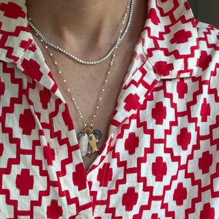 A close-up of a person wearing a red and white geometric patterned button-up shirt with layered silver necklaces and light blue denim jeans.