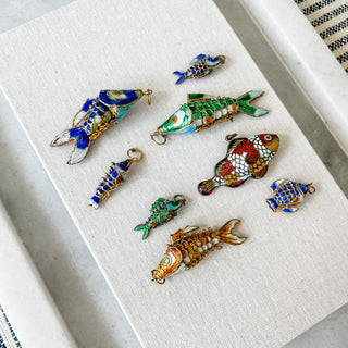 A set of nine colorful fish-shaped jewelry charms or pendants arranged on a textured white fabric surface. The fish designs feature intricate enamel detailing in blue, green, red, orange, and gold, with some pieces showing scales and others displaying abstract patterns.