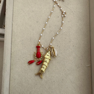 A gold chain necklace with white pearl-like beads, featuring a red lobster and fish charm, displayed inside a beige jewelry box.