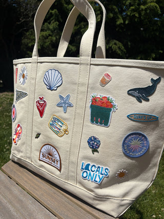 Beige tote bag with various embroidered patches on a wooden surface.