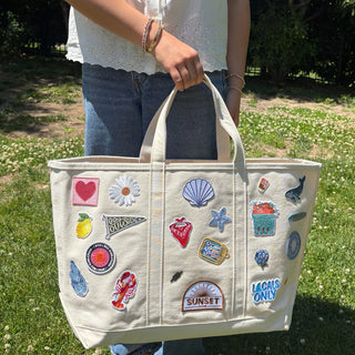 Beige tote bag with various patches held by a person outdoors.