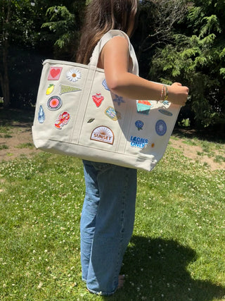 Person holding a beige tote bag with colorful patches in a grassy outdoor setting