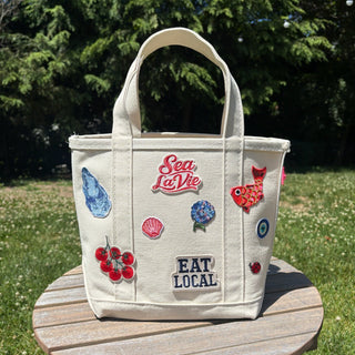 Beige tote bag with colorful patches and text on a wooden table outdoors.