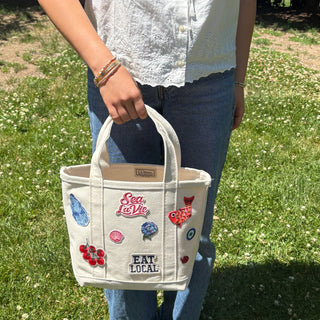 Person holding a white tote bag with colorful designs and text in an outdoor setting.