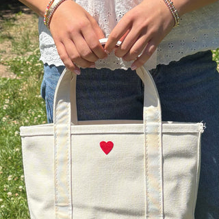 Person holding a white tote bag with a red heart on grass