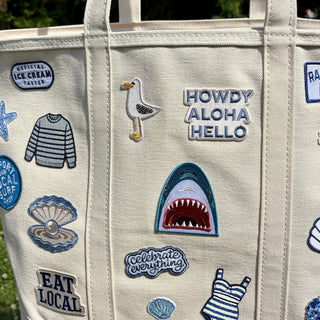 Beige tote bag with various patches including a shark, seagull, and text phrases.