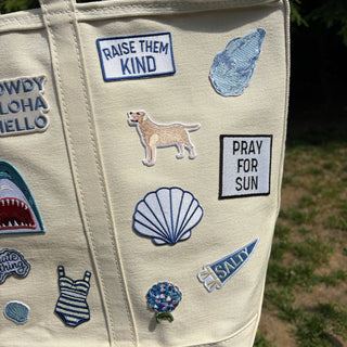 Beige tote bag with various patches including a dog, seashell, and text phrases.