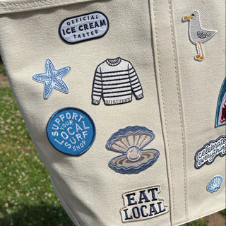 Beige tote bag with various patches including a starfish, ice cream truck, and 'Eat Local' text.