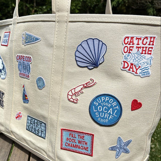 Beige tote bag with various colorful patches on a wooden surface outdoors.