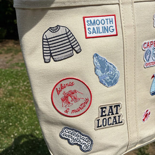 Beige tote bag with various patches including 'Smooth Sailing', 'Eat Local', and others.